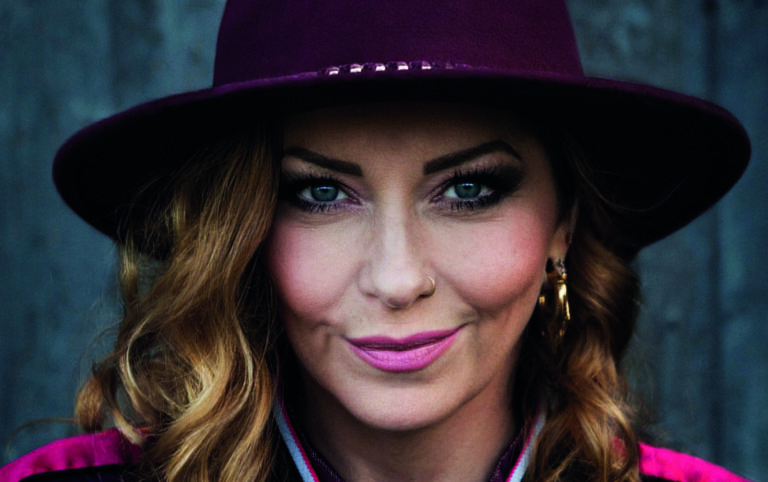 Een portretfoto van Anneke van Giesbergen met een paarse jas aan en een paarse hoed op.