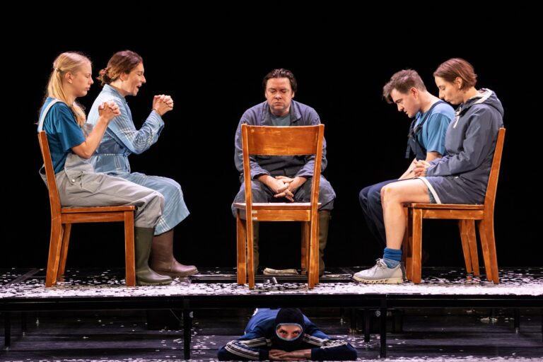 Een theatrale scène met vijf acteurs, waarvan vier zittend op stoelen en één liggend op de grond, gekleed in verschillende kostuums.