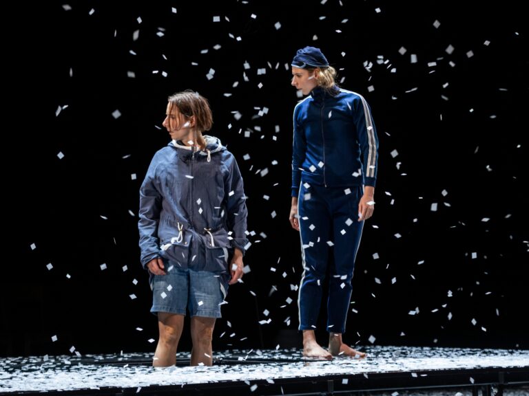 Twee performers staan op een podium met neerdalende sneeuwvlokken tegen een donkere achtergrond.