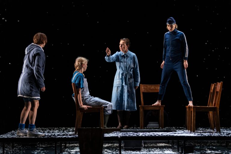 Een toneelstuk met vier acteurs in een donkere setting, waarbij één persoon een boodschap overbrengt terwijl de anderen op stoelen staan of zitten.