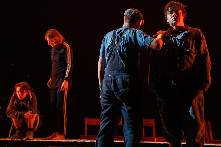 Drie acteurs staan op het podium in een donkere setting, met een focus op hun interactie en emoties.
