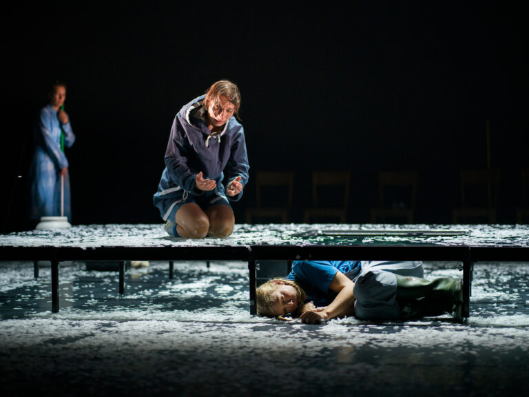 Twee acteurs op een podium bedekt met papieren snippers, waarbij één op de grond ligt en de ander knielend lijkt te expressieve emoties te tonen.
