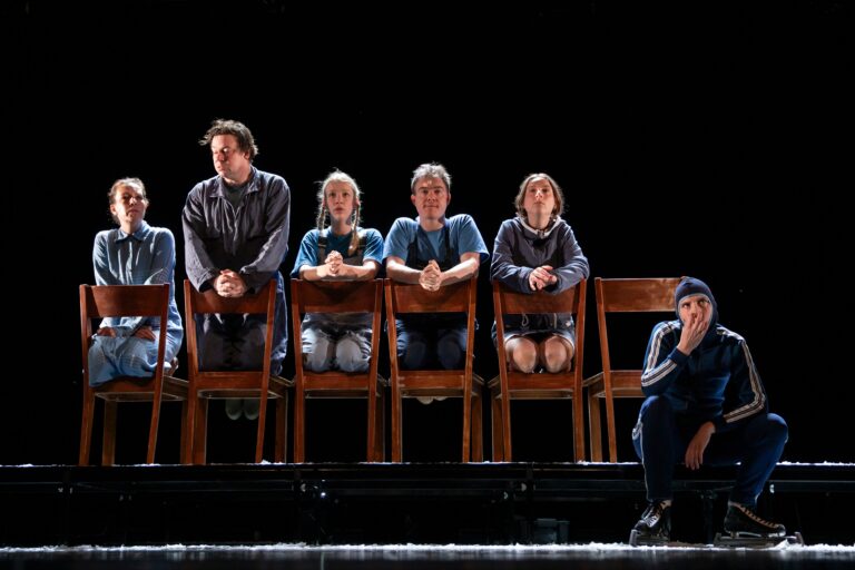 Een groep acteurs zit op stoelen op een podium met een donkere achtergrond.