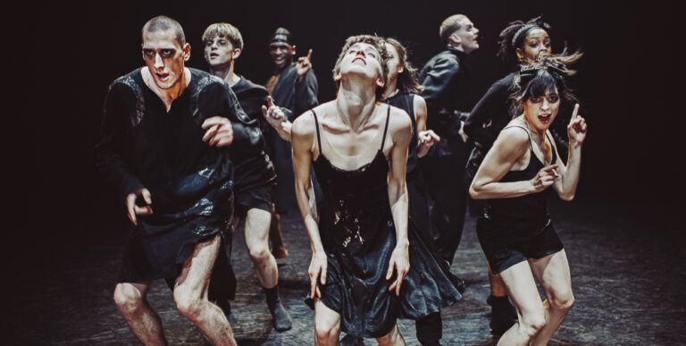 Dansers in zwarte kleding staan met de ruggen naar elkaar toe in een cirkel.