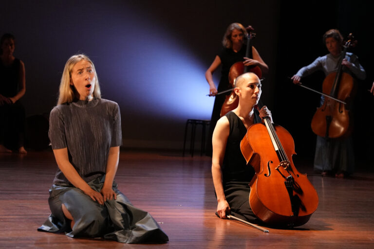 Twee personen die op hun knieën zitten. De één zingt de ander heeft een cello in de hand.