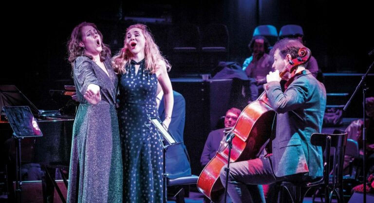 Drie zangeressen optreden op een podium, terwijl een cellist hen begeleidt.