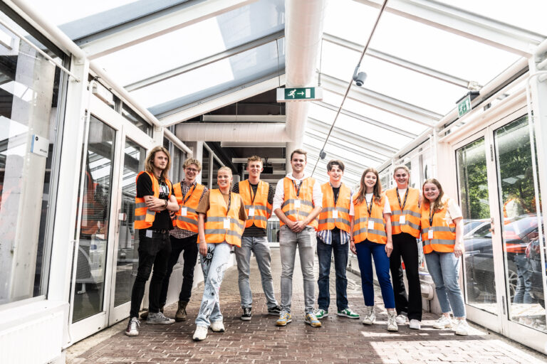 Een groep van negen mensen in oranje veiligheidshesjes poseert in een glazen hal.