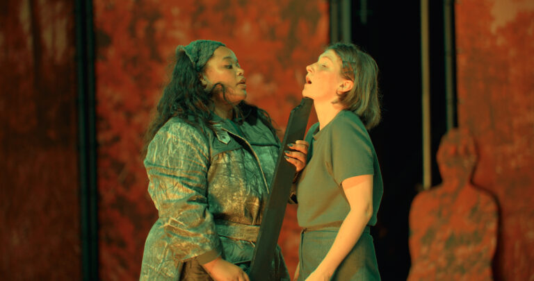 Scènefoto van twee actrices die tegenover elkaar staan. Een actrice houdt een geweer onder het gezicht van de ander.