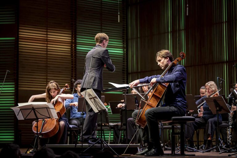 Een klein orkest speelt muziek, met een dirigent die aanwijzingen geeft en een cellist die zijn partituren leest.