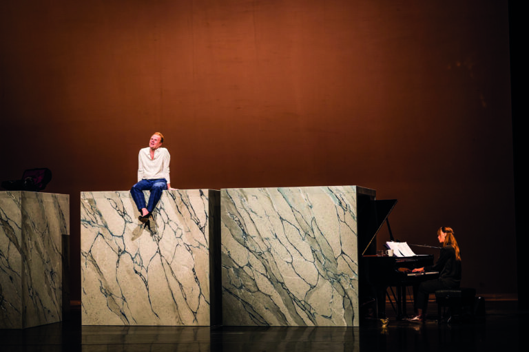 Een acteur zit op een grote marmeren blok, terwijl een pianist aan de rechterzijde van het podium speelt.