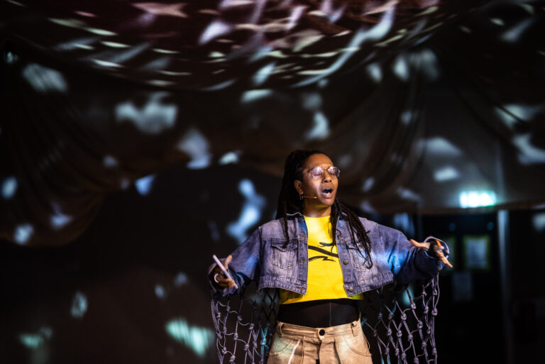 Een persoon in een sprekende houding, gekleed in een gele tee en spijkerjacket, staat voor een achtergrond met kleurrijke lichtprojecties.