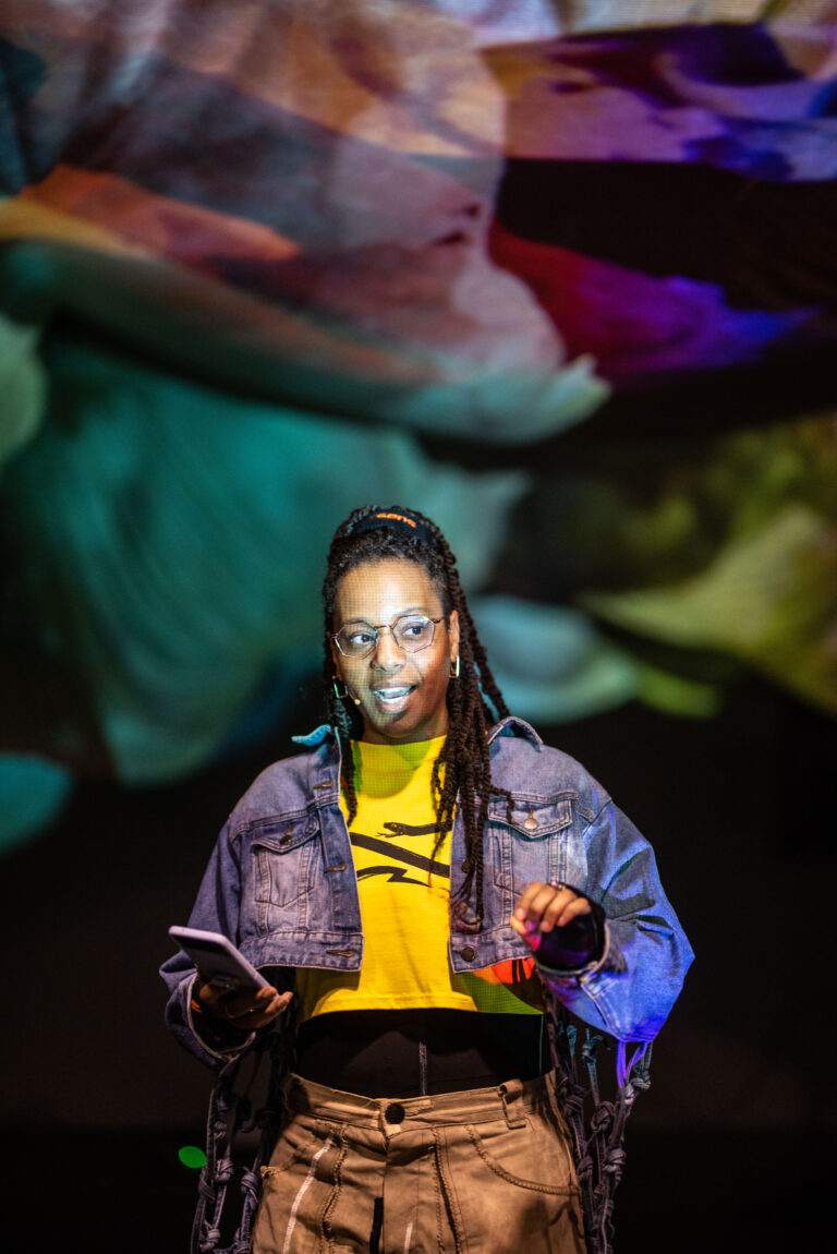 Een persoon in een spijkerjack en gele sweater staat op een podium met een kleurrijke achtergrond.