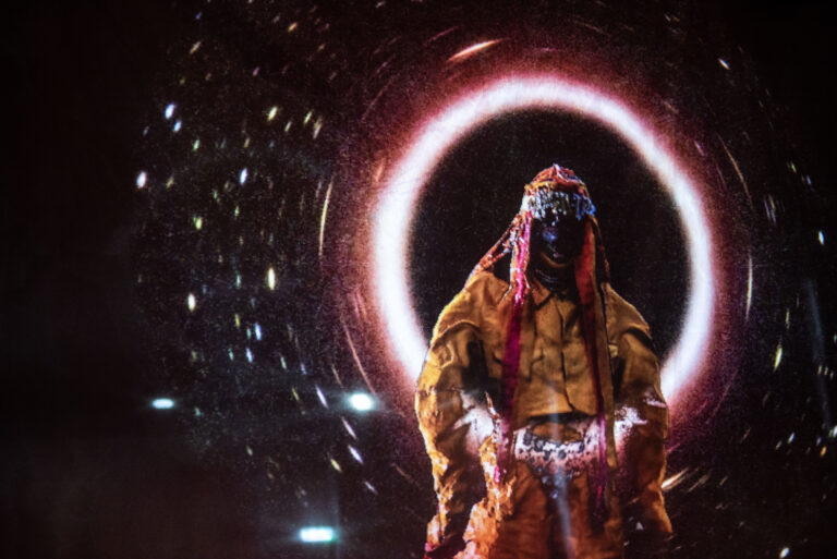 Een persoon in een kleurrijke kostuum staat met een mysterieuze, lichtgevende cirkel op de achtergrond.