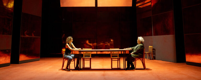 Twee acteurs zitten tegenover elkaar aan een lange tafel