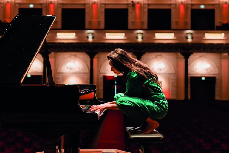 Een muzikante in een groene outfit speelt piano in een sfeervolle concertzaal.