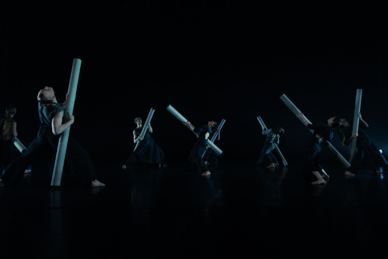 Groep dansers in donkere ruimte die met lange stokken bewegen.