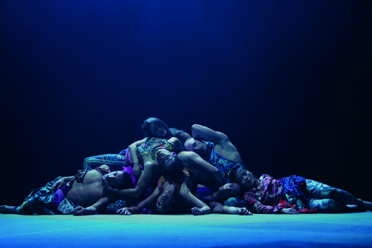 Een groep mensen ligt op elkaar gestapeld op een podium onder een blauwe verlichting.