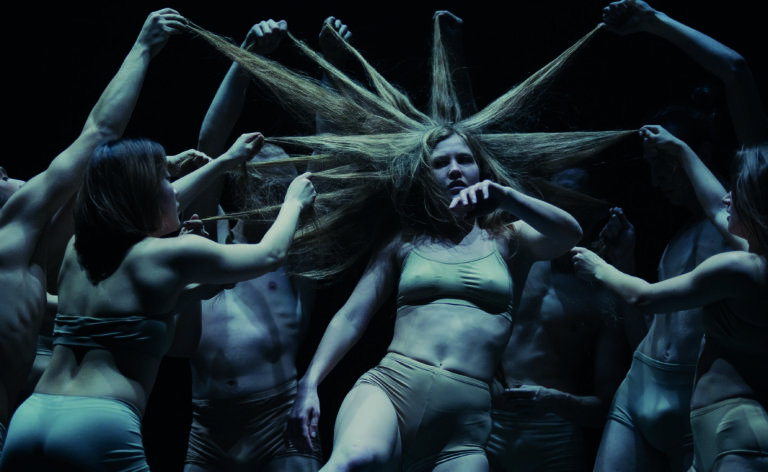 Een krachtige dansperformance met een centrale figuur omringd door andere dansers, die allemaal op unieke wijze hun lange haar gebruiken.