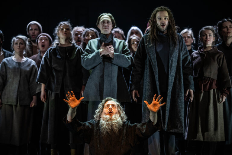 Zingende mannen in donkere gewaden. Op de voorgrond een man met lang grijs haar en een lange grijze baard.