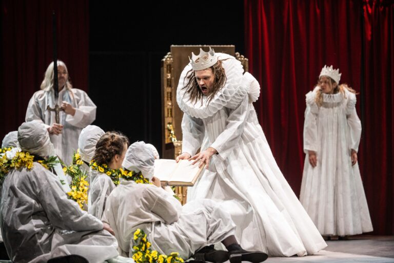 Zittende mensen in witte pakken en een gele bloemen ketting. In het midden een man met lang haar en een baard met een witte kroon.