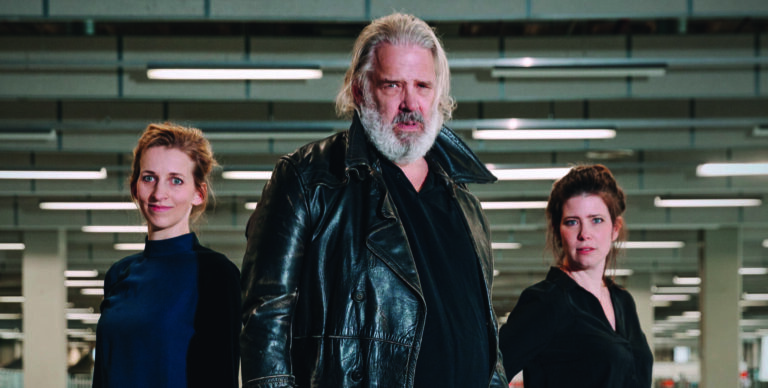 Drie mensen staan in een industriële omgeving, met een focus op een man in het midden met een lange leren jas en grijs haar.