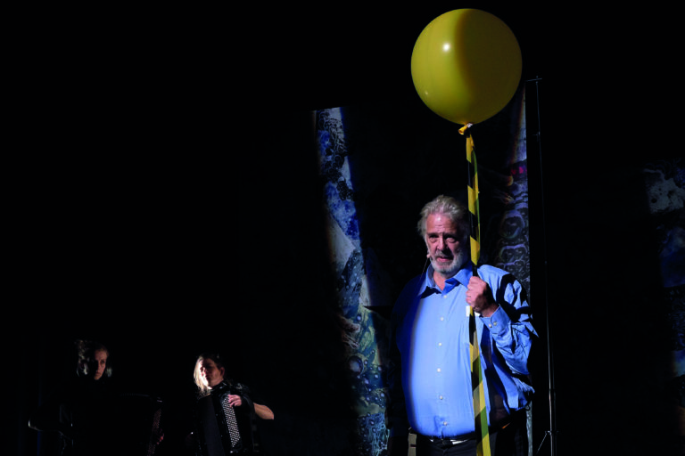 Een man met een gele ballon staat in een donkere omgeving, terwijl een andere persoon op de achtergrond te zien is.