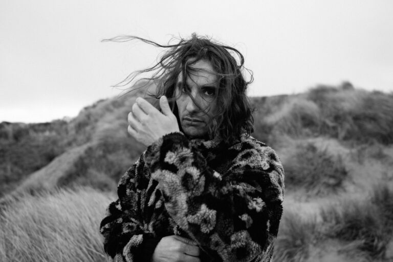 Een man met lang, warrig haar staat in een veld, terwijl hij zijn hand naar zijn gezicht brengt, gekleed in een fluffy, bloemenpatroon jas.