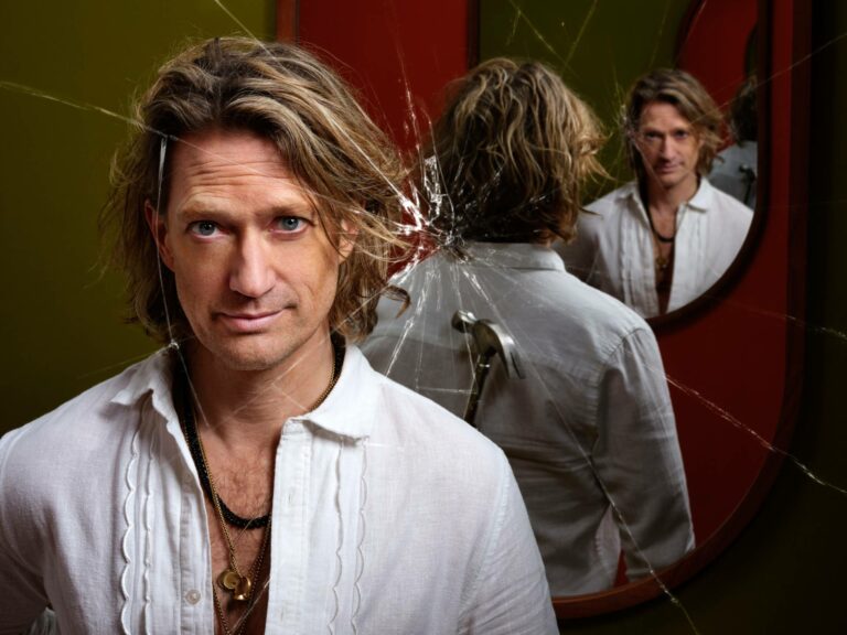 Joost Spijkers die in een kapotte spiegel staart terwijl hij een hamer achter zijn rug houdt. Hij draagt een wit overhemd en veel gouden kettingen.