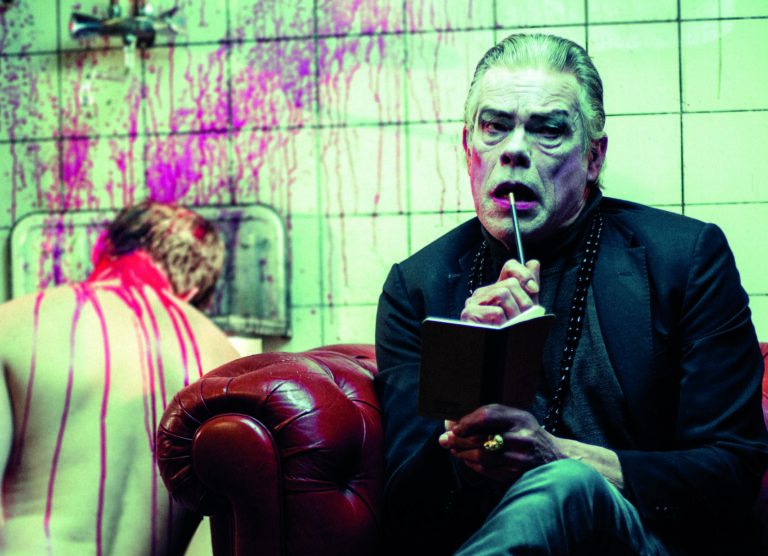 Een man met een clownachtig uiterlijk zit op een leren bank en schrijft in een notitieboekje, terwijl er achter hem een omhulde figuur met bloedsporen op de muur zichtbaar is.