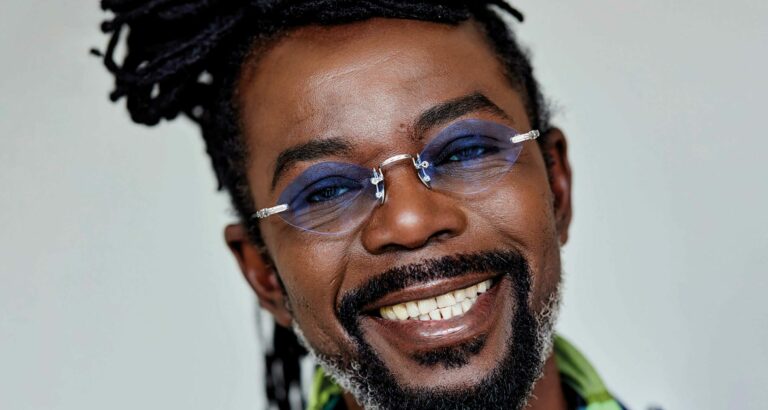 Close up foto van zanger Kenny B voor een grijze muur, hij kijkt vrolijk naar de camera.