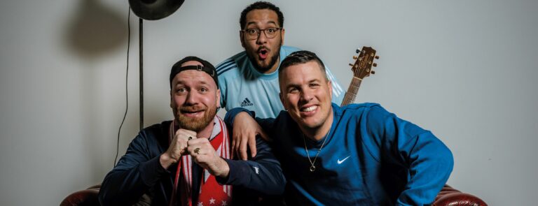 Drie mannen poseren vrolijk samen, met een gitaar zichtbaar op de achtergrond.