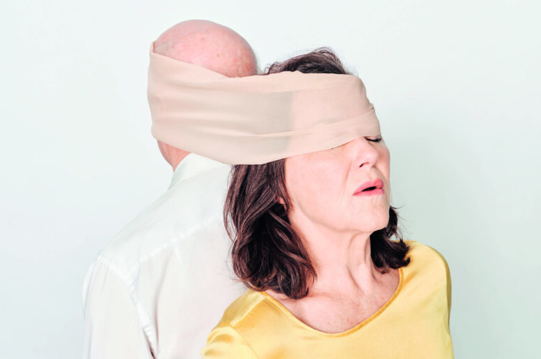 Acteur en Actrice met de ruggen tegen elkaar aan en geblinddoekt met 1 groot doek.