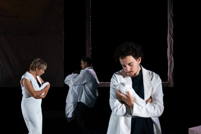 Drie mensen in labjassen houden een baby of een object dicht bij zich terwijl ze in een donkere omgeving staan.