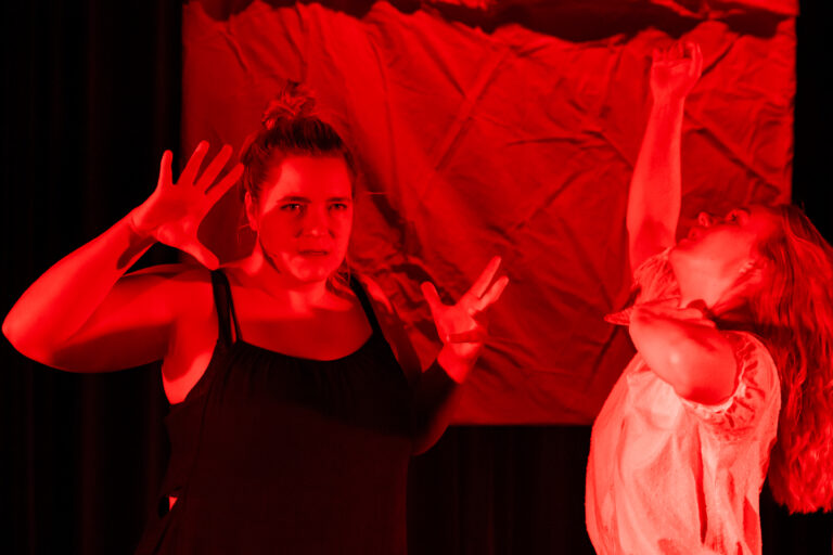 Twee vrouwen voeren een expressieve performance uit onder een rode belichting.