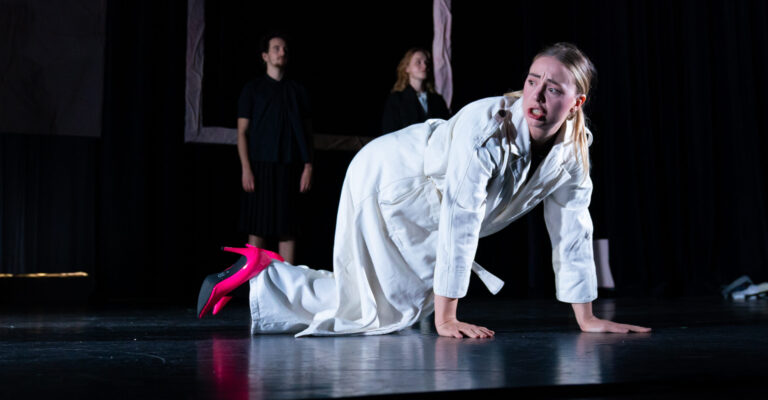 Actrice op haar handen en knieën op het podium in een wit pak met roze hakken.