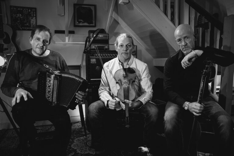 Drie muzikanten zitten in een kamer, met verschillende instrumenten in hun handen.