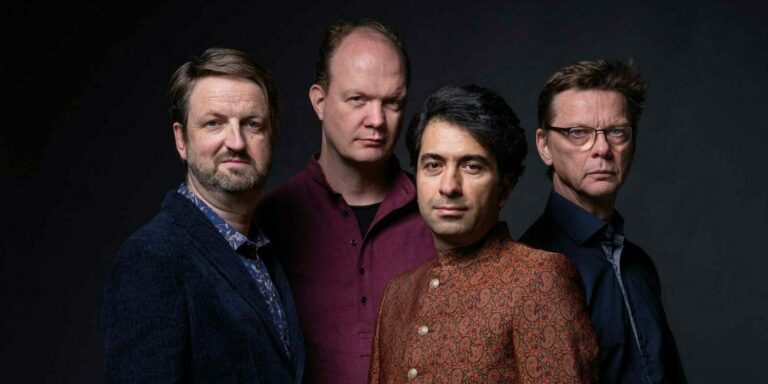 Een groep van vier mannen poseert in een donkere achtergrond, ieder in verschillende kledingstijlen.