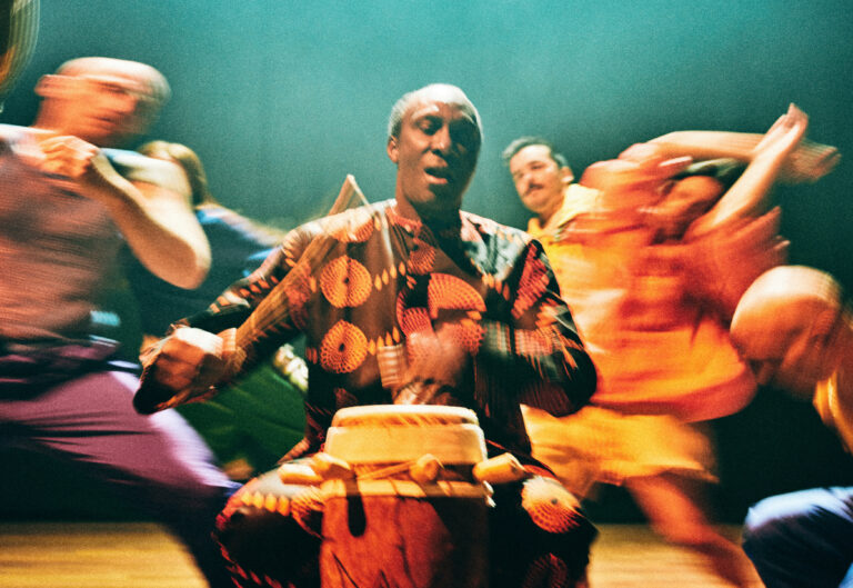 Een muzikant in traditionele kleding speelt op een djembé, omringd door dansers in beweging.