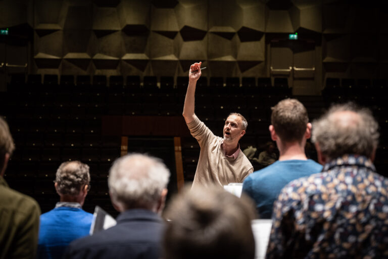 De dirigent, een man heeft een licht shirt aan en een hand in de lucht