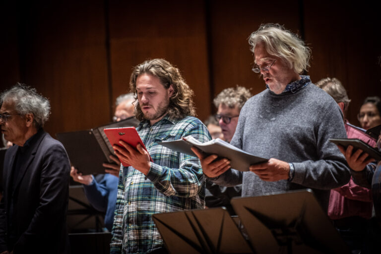 Twee mannen in een grijze trui en geblokte blouse kijken naar hun bladmuziek en zingen