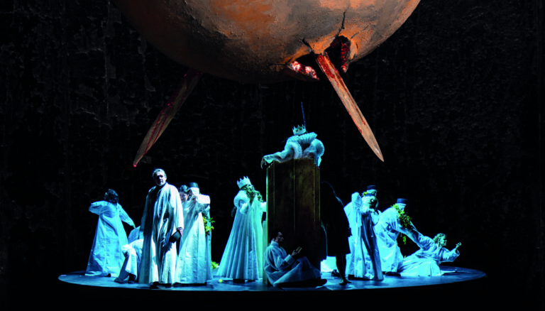 Dansende, verklede mensen in witte kleding in blauw licht. In de lucht hangt een rond voorwerp met daar doorheen een punt.