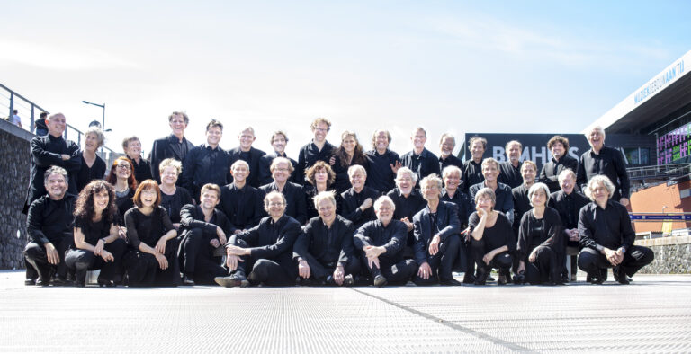 Groepsfoto van een ensemble in zwarte kleding, poserend buiten met een blauwe lucht op de achtergrond.