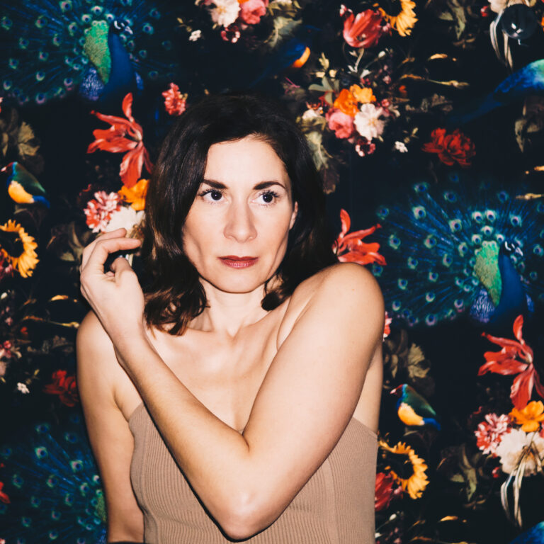 Een vrouw met een neutrale uitdrukking poseert voor een kleurrijke, bloeiende achtergrond.