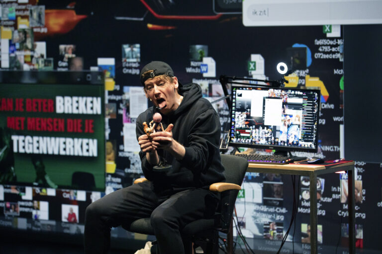 Een man in een zwart sweatshirt zit op een stoel en spreekt enthousiast in een microfoon, met een achtergrond van beeldschermen en gekleurde graphics.