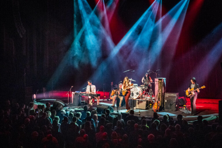 Een band speelt live op een podium met kleurrijke lichtstralen en een enthousiast publiek.