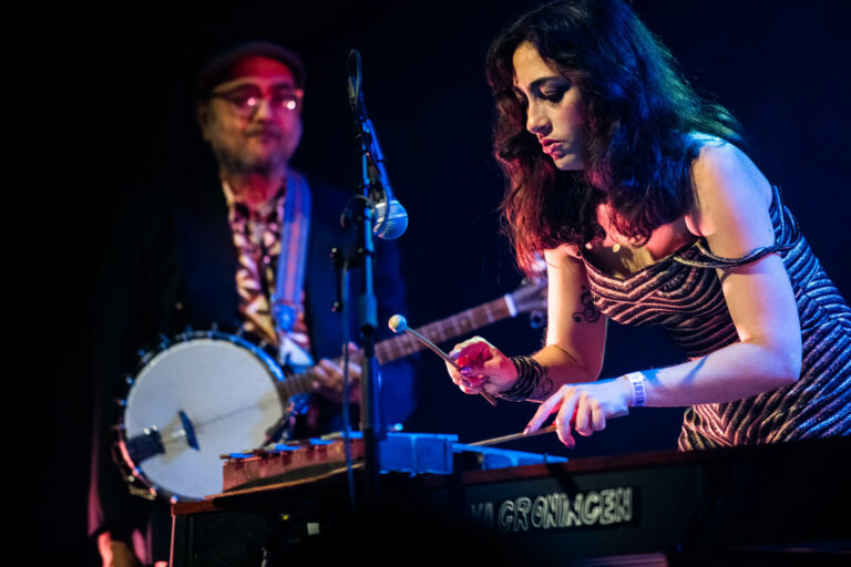 Een muzikale optredens met een vrouwelijke muzikant die een instrument bespeelt, terwijl een muzikant met een banjo op de achtergrond speelt.