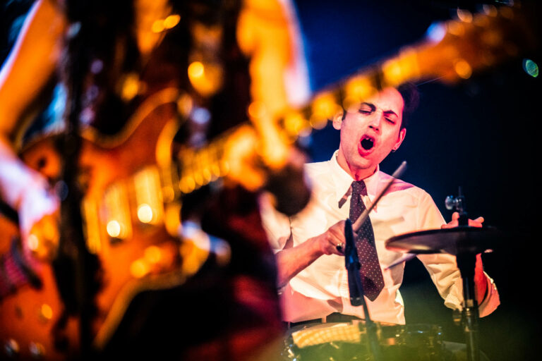 Een drummer speelt intens op een podium, terwijl een gitarist deels onscherp op de voorgrond zichtbaar is.