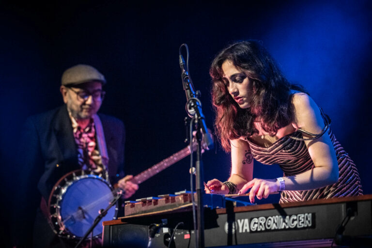 Een muzikante speelt op een keyboard terwijl een man met een banjo op de achtergrond staat, beide in een sfeervolle, verlichte setting.