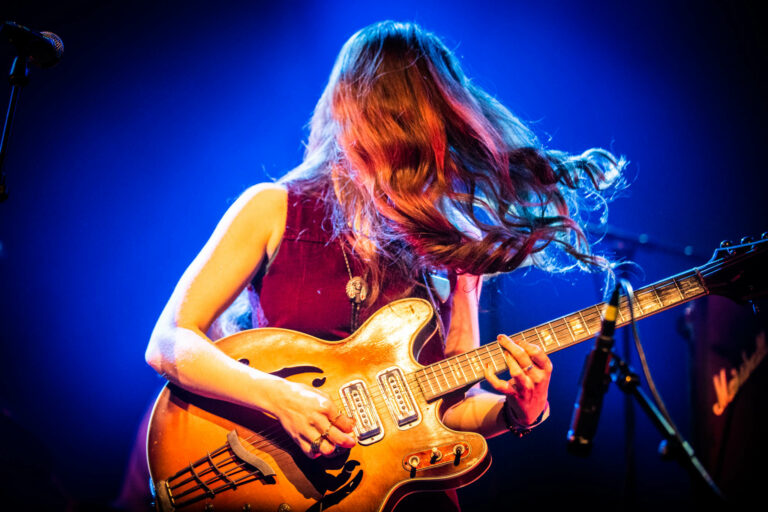 Een muzikante met lang, golvend haar speelt een elektrische gitaar op een podium met blauwe verlichting.