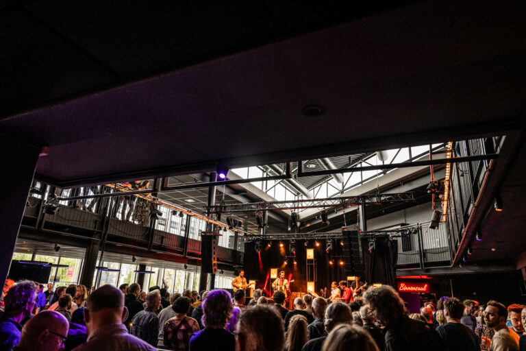 Een drukbezocht concert met een band die optreedt in een grote, industrieel ogende locatie.